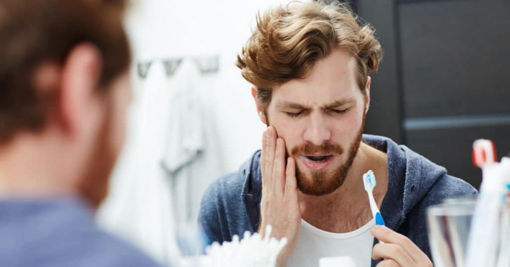 Le reflet d'un homme dans un miroir : il tient sa brosse à dent d'une main et de l'autre se tient la mâchoire, visiblement douloureuse.