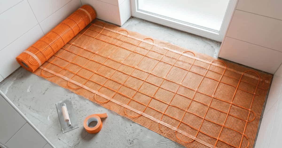 Tapis chauffant électrique orange partiellement déroulé sur une chape brute dans une salle de bains carrelée de blanc avec une spatule et un rouleau de ruban adhésif posés à côté.