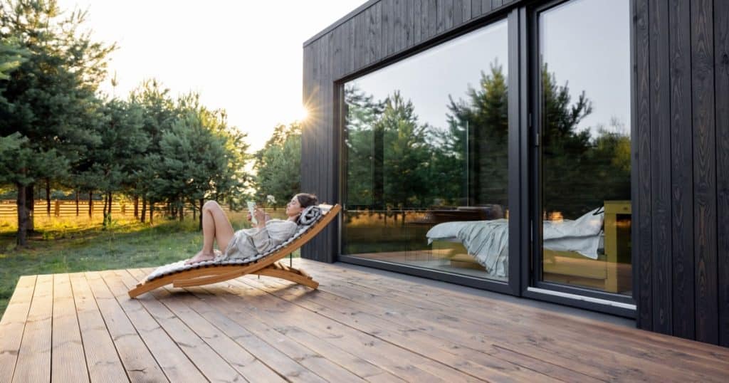 terrasse en bois clair donnant sur un jardin boisé avec une femme en train de lire un livre au soleil sur un transat