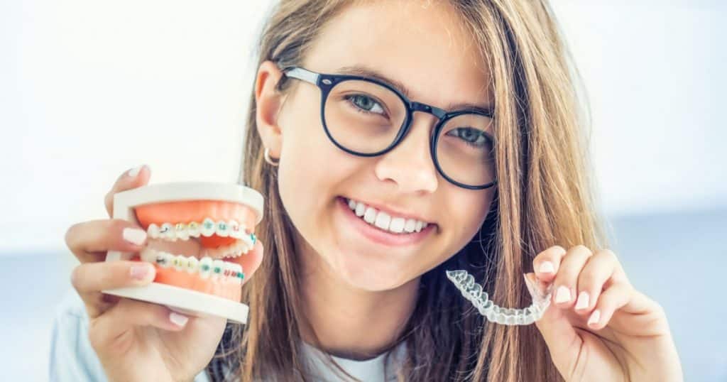 Une adolescente montre un sourire parfait en tenant d'une main une maquette de dentition avec un appareil dentaire et de l'autre une gouttière d'alignement transparente. 