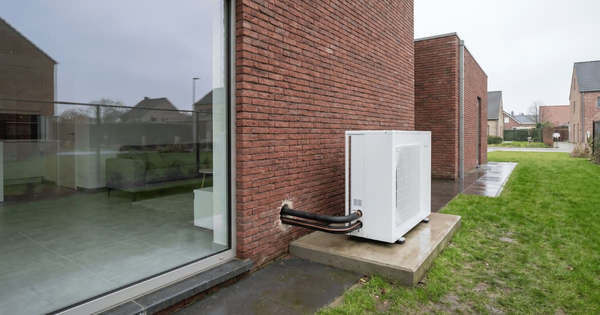 Unité extérieure blanche d'une pompe à chaleur installée sur un socle en béton contre une façade en brique rouge d'une maison moderne dans un quartier résidentiel belge par temps couvert.