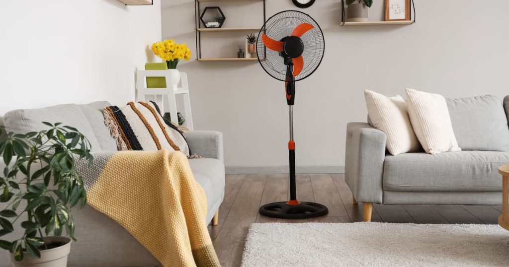 Une chambre sans climatisation que l’on souhaite rafraîchir à l’aide d’un ventilateur. La pièce contient un lit et une armoire ; deux tapis sont posés sur le sol en carrelage. Le mur, dans une teinte bleu-gris clair, est décoré d’étagères murales blanches. Plusieurs plantes d’intérieur y sont disposées, contribuant également à une température agréable pour le sommeil.