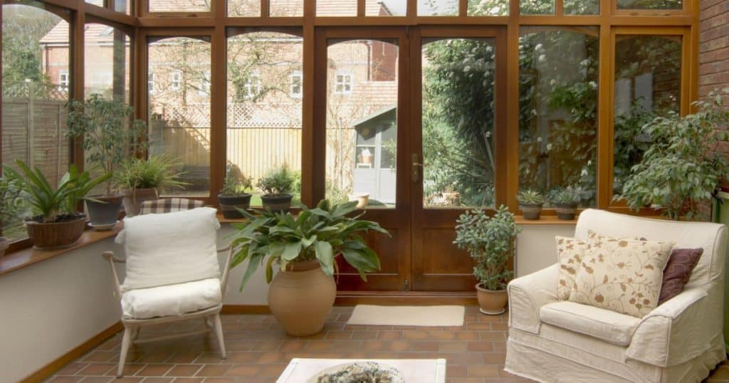 veranda vitrée avec structure en bois abritant des plantes et deux fauteils blancs