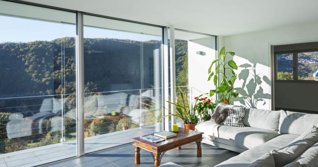 Une véranda avec des stores sur les baies vitrées, une vue sur la nature, et un coin salon aménagé avec une table, des plantes vertes et un canapé. 