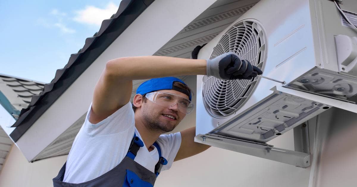 Een erkende airco-installateur monteert de buitenunit aan een huis. Hij draagt een wit shirt met een blauwe overall en een blauwe pet met een veiligheidsbril.