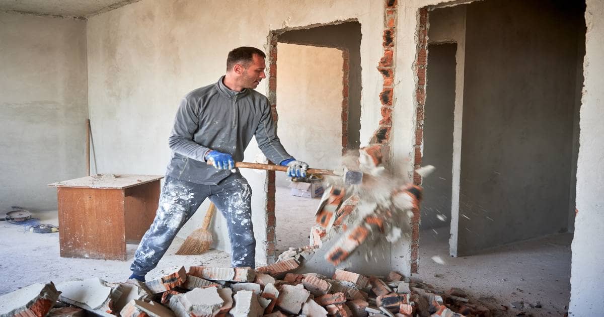 Een man gebruikt een sloophamer om een muur van een woning te slopen. Er vliegen stukken baksteen op de grond.