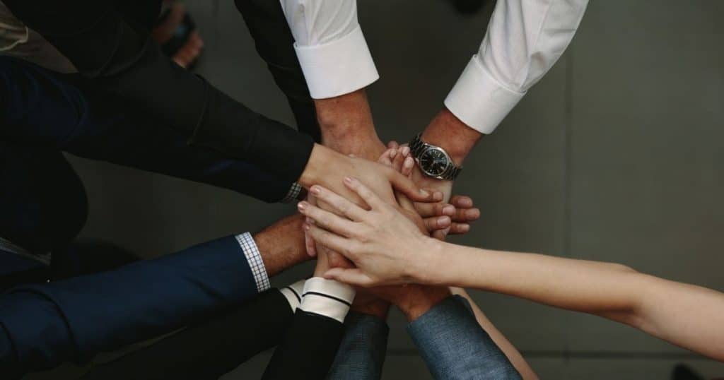 Verschillende mensen staan in een cirkel en leggen de handen op elkaar. Enkel hun handen, armen en mouwen zijn zichtbaar. Ze staan op een vloer met grote, lichte tegels.
