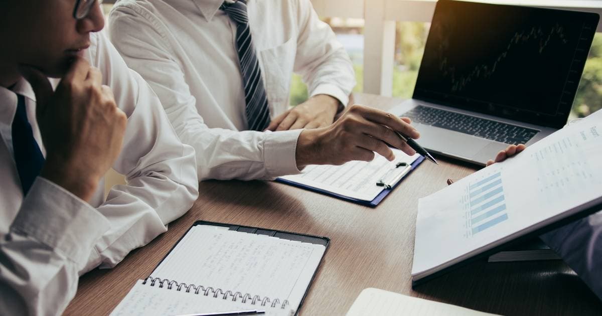 Drie personen zitten aan een houten bureautafel. Achteraan op de tafel staat een laptop. Er liggen ook een klembord en notitieboek op de tafel. De achterste man draagt een wit hemd en een grijs met zwart gestreepte das. Hij wijst iets aan op een document met grafieken, dat vastgehouden wordt door een persoon van wie enkel de hand zichtbaar is. De voorste man strijkt nadenkend over zijn kin en draagt een bril, wit hemd en een das.