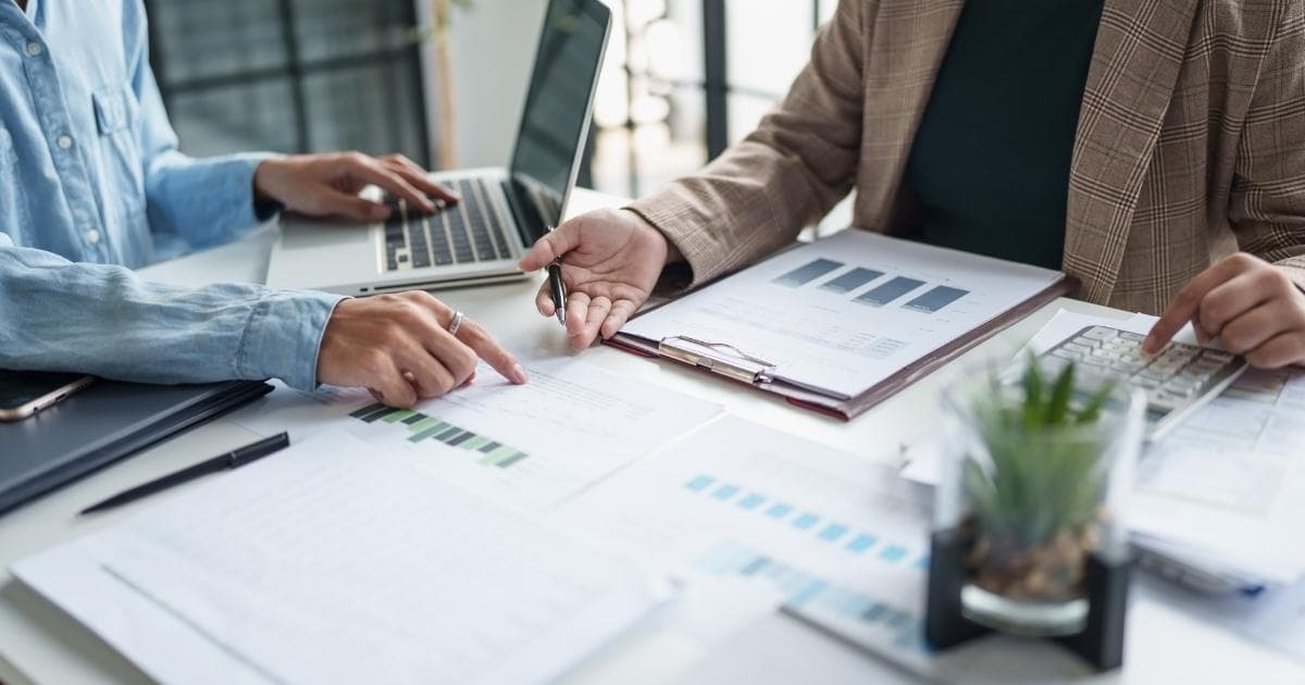 De bovenlichamen van twee personen aan een grijze tafel. Er liggen verschillende documenten met grafieken en tabellen op tafel. De linkse persoon draagt een lichtblauw hemd met witte knoopjes. De persoon wijst iets aan op één van de documenten met de rechterhand, terwijl de linkerhand op het toetsenbord van een grijze laptop rust. De rechtse persoon draagt een zwart shirt onder een bruine vest met ruiten. De rechtse persoon heeft de linkerhand boven een rekenmachine, die op een stapel papieren ligt. In de rechterhand houdt de persoon een pen vast. De handpalm van de rechterhand wijst naar boven. Voor hen, in de hoek rechtsonder, staat een vetplantje in een glazen potje dat in een zwart houdertje zit.