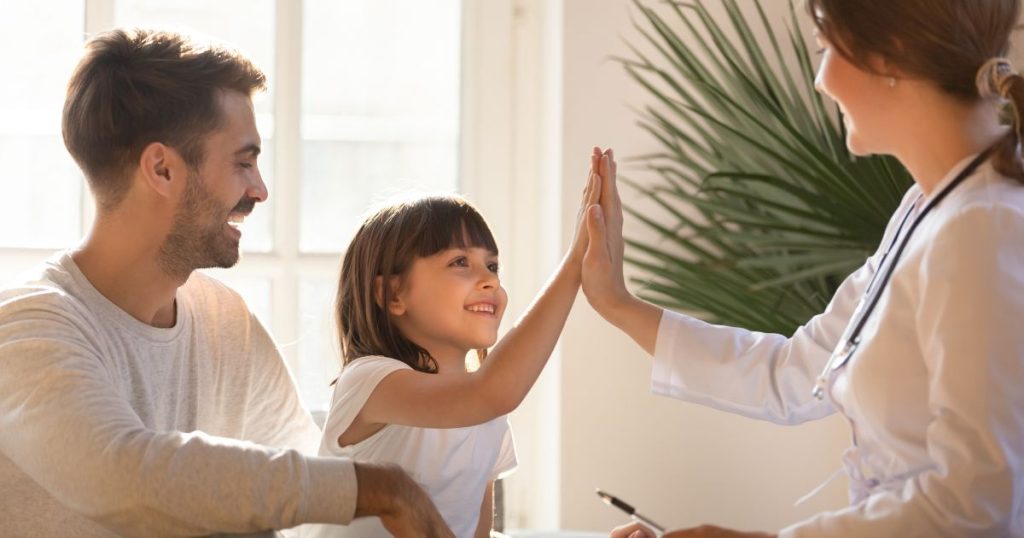 Lachende arts geeft high five aan meisje, vader kijkt toe tijdens consult over aanvullende ziekteverzekering