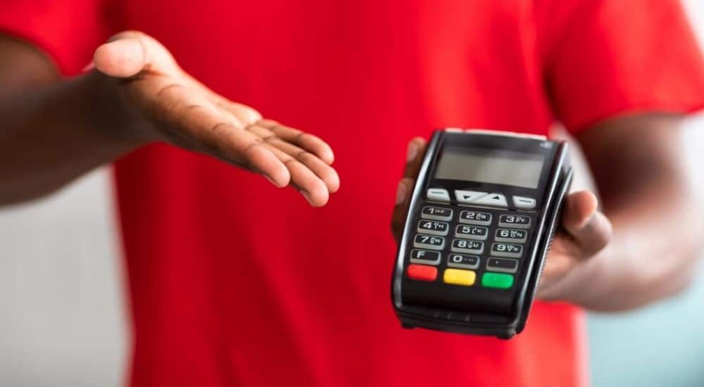 Een man met een rood t-shirt gebaart naar een betaalterminal in zijn hand. Die betaalterminal maakt het mogelijk om met een bankkaart te betalen.