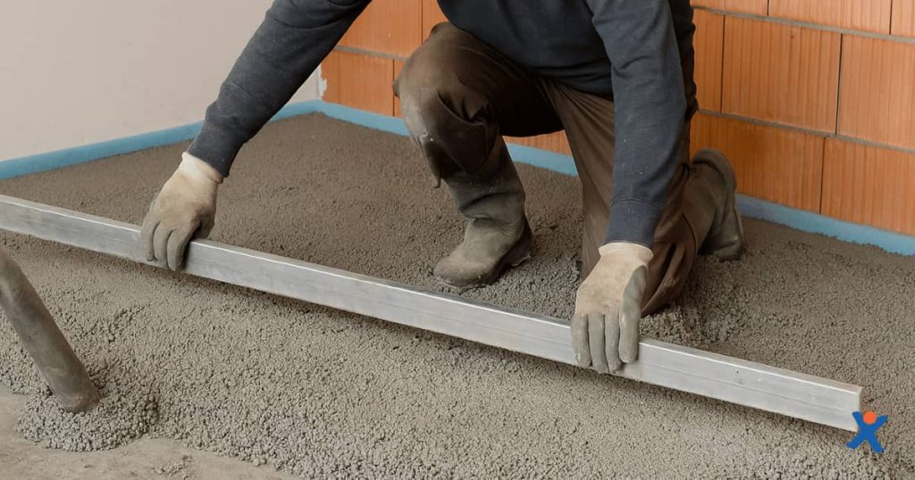 Een chapper spreidt isolerende chape gelijkmatig over een vloer in een onafgewerkte ruimte met een metalen rei, terwijl het materiaal met kleine korrels een egale laag vormt.