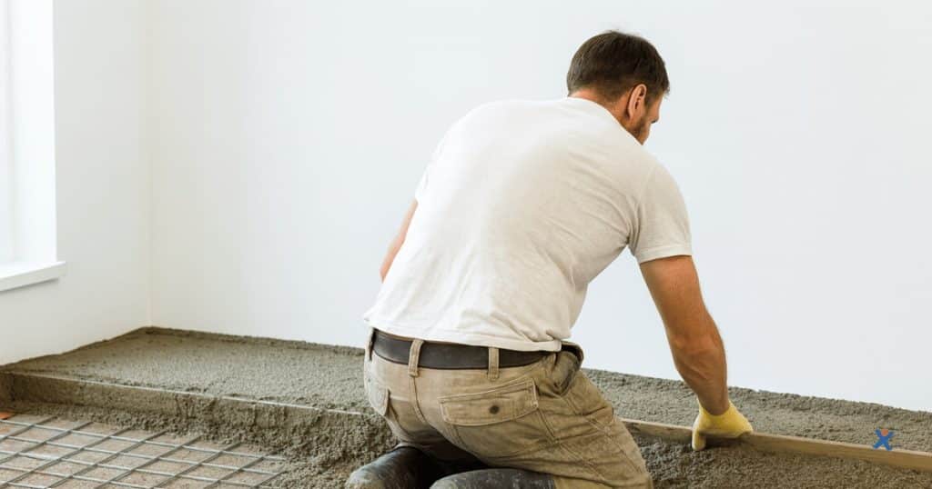 Vakman op knieën die gewapende chape over vloerverwarming en metalen netten gladstrijkt met een rei in een lichte kamer.