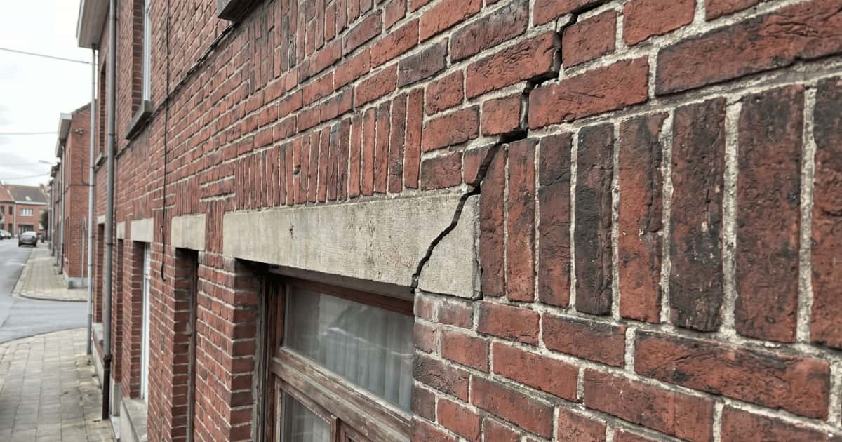 Een brede scheur loopt vanuit een betonnen latei boven een raam door het metselwerk naar boven in de gevel van een Vlaamse rijwoning.