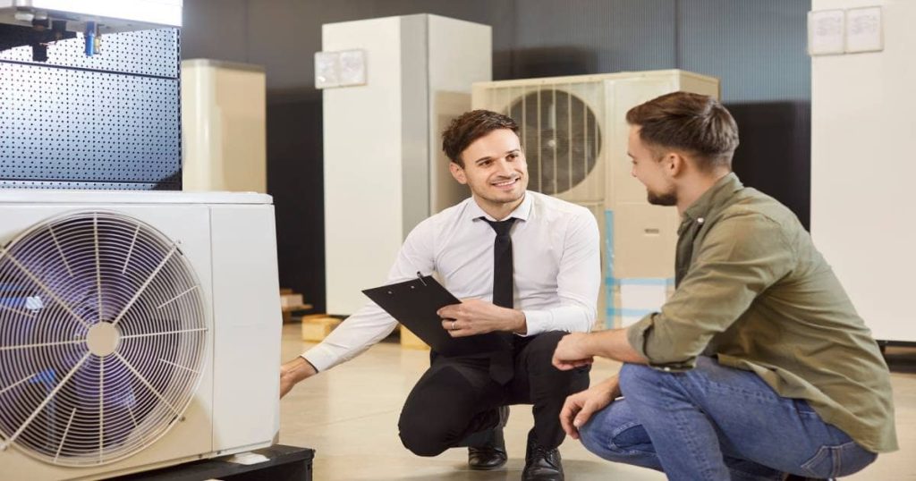 Een verkoper met wit hemd en zwarte das wijst met zijn hand naar een airco in een showroom. Samen met een andere man zitten ze gehurkt bij het betreffende toestel.