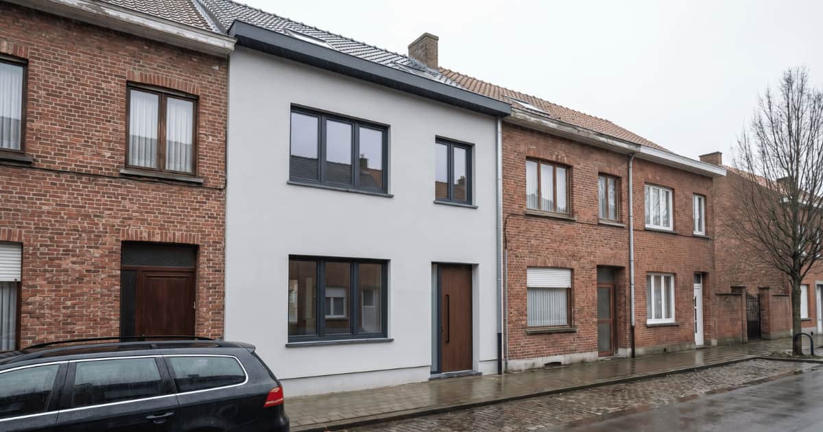 Vlaamse rijwoning met strakke witte crepi-afwerking en donkere raamprofielen, opvallend contrast met de ongerenoveerde bakstenen buurwoningen op een regenachtige dag.