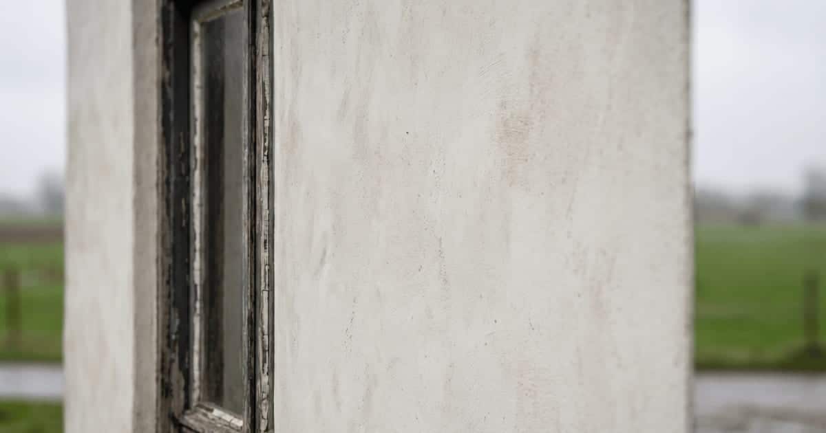 Kalkpleister op een Vlaamse gevel toont de typische matte en licht onregelmatige minerale afwerking met subtiele streken naast een verweerd raamkozijn tegen een groen landelijk decor.