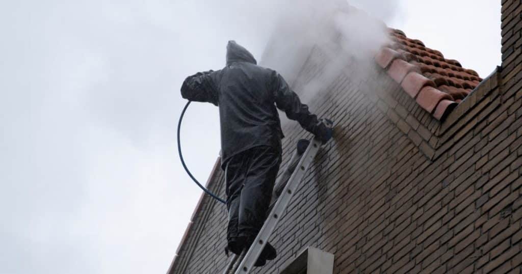 Specialist reinigt bakstenen gevel met stoom op ladder bij nat weer.