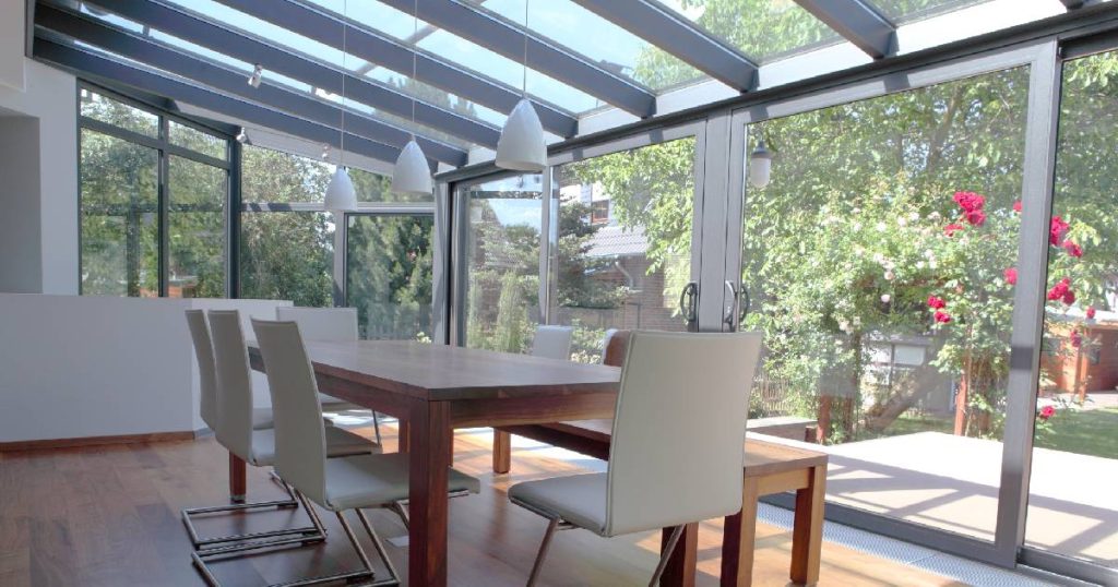 Een veranda in dubbel glas met aluminium profielen van Horrix veranda's, een bedrijf met positieve reviews. In de veranda staat een houten tafel, houten zitbank en 3 moderne witte stoelen met ijzeren poten.