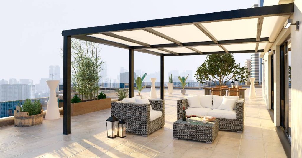 Een aluminium pergola met semi-transparant dak uit dubbel glas op een hooggelegen terras, zoals ook wordt geplaatst door Horrix veranda's, een bedrijf met positieve reviews. Onder de pergola staat een rieten loungeset met witte kussens. 