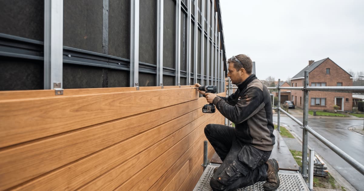 Een schrijnwerker bevestigt horizontale houten gevelplanken met een accuboormachine op een metalen onderconstructie terwijl op de achtergrond een Vlaamse bakstenen woonwijk zichtbaar is op een bewolkte dag.
