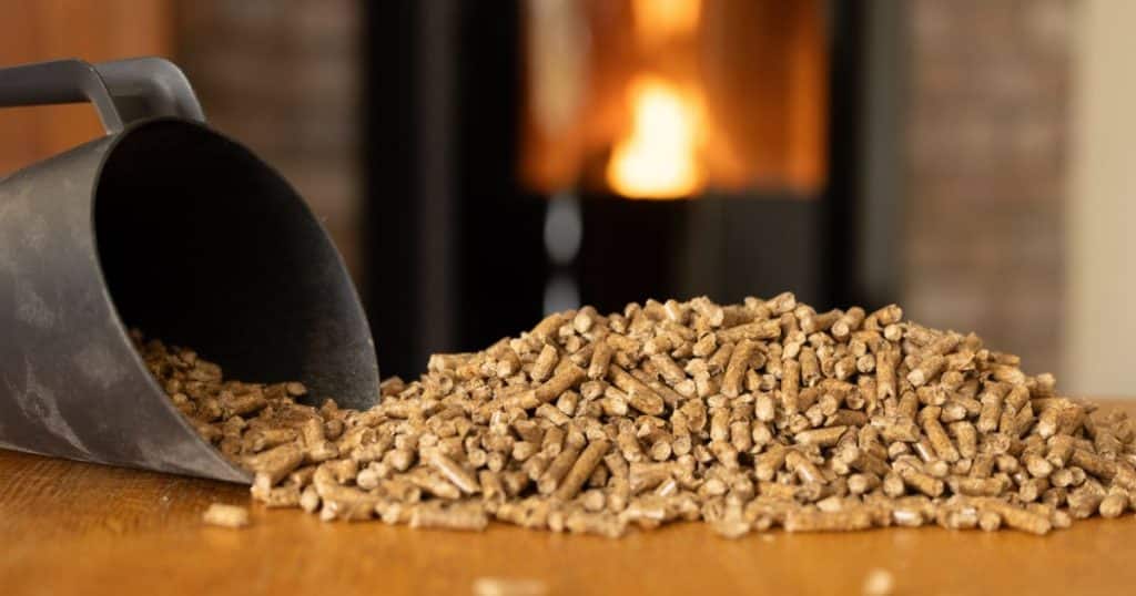 Schep met houtpellets op een houten tafel, met een brandende pelletkachel op de achtergrond