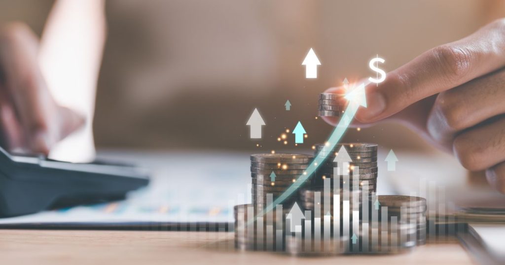 Een hand legt enkele muntstukken op een stapel. Op de achtergrond typt hij vluchtig iets in op een toestel. Een denkbeeldige pijl gaat omhoog en wijst als het ware naar een dollar-teken