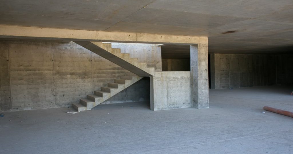 Trap in beton die uitkomt op een grote kelder- of garageruimte. De trap is in 2 delen, met een bordes tussenin.