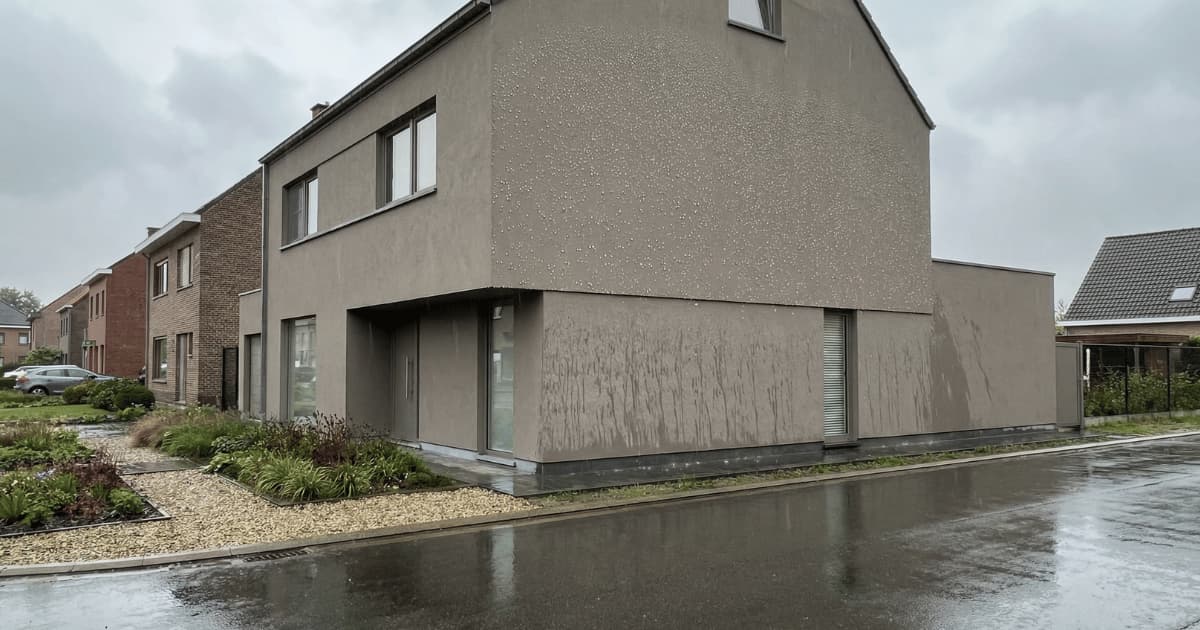 Regendruppels parelen af op de donkergrijze gevelschildering van een moderne Vlaamse hoekwoning terwijl waterstrepen op de benedenverdieping het waterafstotend effect van siliconenverf zichtbaar maken.