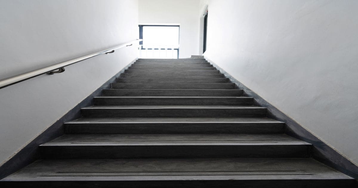 Onderaanzicht van een zwarte trap met betonlook, metalen trapleuning en antislip strips in een witte trappenhal.