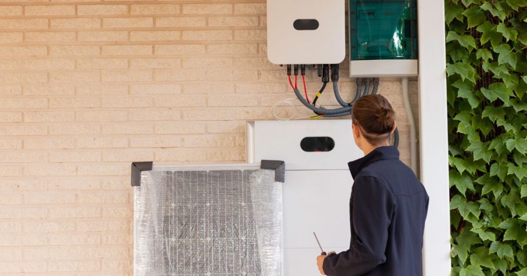 Een installateur in donkerblauwe overall controleert een thuisbatterij installatie. Tegen de installatie rust ook nog een zonnepaneel. Zal de installateur die straks ook nog plaatsen?