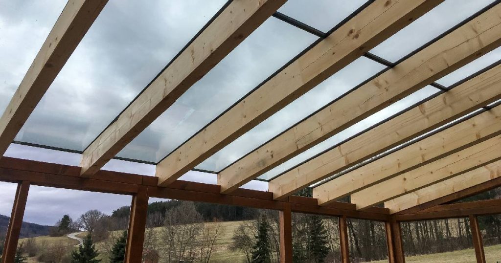 Onderaanzicht van een veranda met glazen dak, ondersteund door houten balken en afgewerkt met screens.