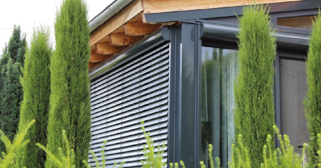 Een glazen veranda met rolluiken, een alternatief op een dak met screens. Voor de veranda staan enkele hoge struiken. Het dak van de veranda is opgebouwd uit hout en voorzien van een dakgoot.