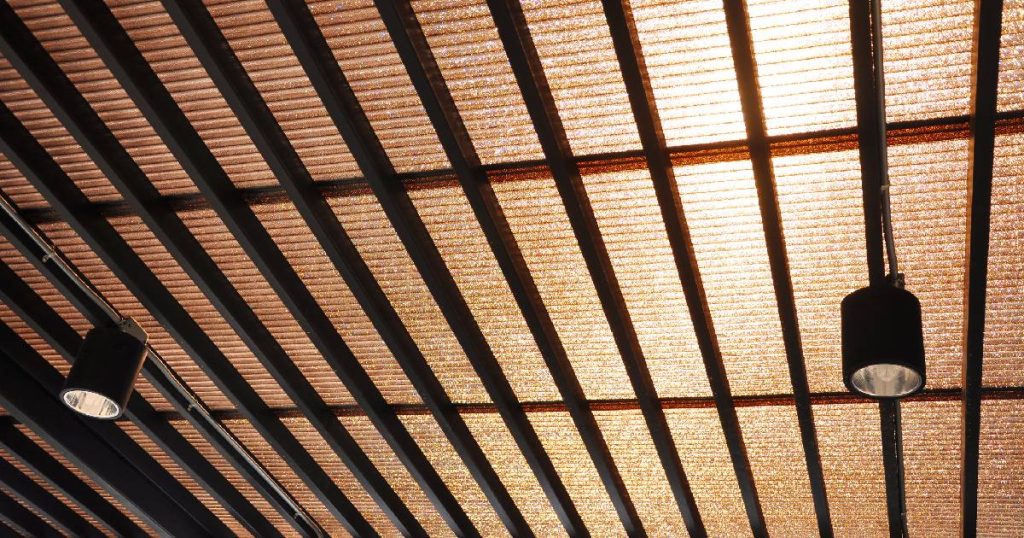 Onderaanzicht van het dak van een veranda uit plexiglas of polycarbonaat, afgewerkt met een screen om hinderlijk zonlicht te remmen. Onder het dak hangen twee zwarte spotjes.