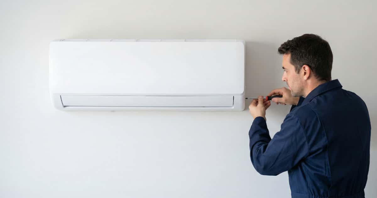 Technicus in donkerblauwe werkkleding die koelleidingen aansluit aan de onderzijde van een witte split airco binnenunit gemonteerd op een lichte muur.