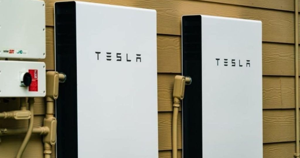 Twee Tesla Powerwalls tegen een lichtbruine, houten wand. Ze zijn beide verbonden met de omvormer aan de linkerkant.