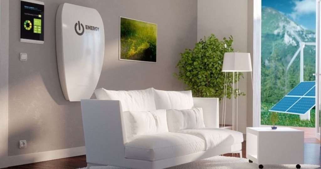 Een grote witte thuisbatterij hangt in een woonkamer met een witte bank. Er hangt een schilderij met een landschap aan de muur en in de hoek van de kamer staat een boompje. Uit het raam zijn bergen, een windturbine en zonnepanelen te zien.