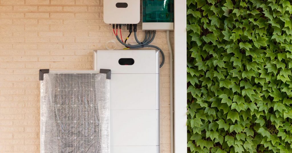 thuisbatterij-installatie tegen een lichte bakstenen muur. Tegen de muur rust ook een zonnepaneel in cellofaan verpakking.