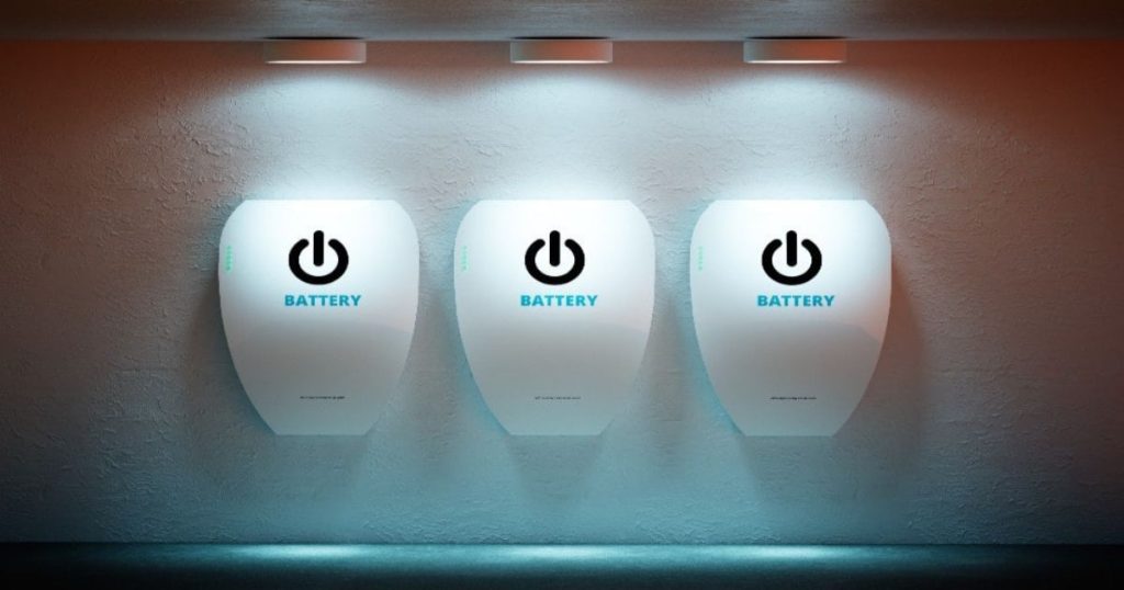 Drie witte thuisbatterijen tegen een witte muur. Ze worden belicht door drie spots aan het plafond.