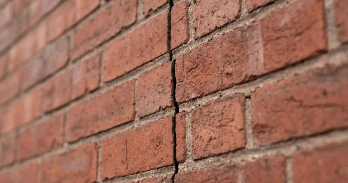 Een verticale scheur loopt recht door meerdere baksteenlagen en voegen in een rode bakstenen muur met lichte opening tussen beide zijden.