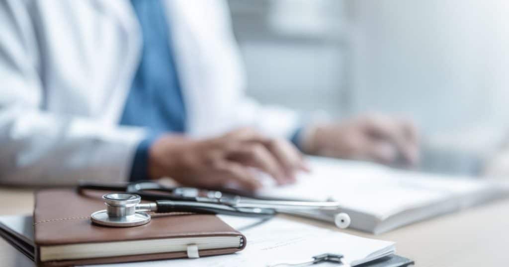 Arts schrijft verslag met stethoscoop op bureau, symbool voor Vlaamse sociale bescherming.