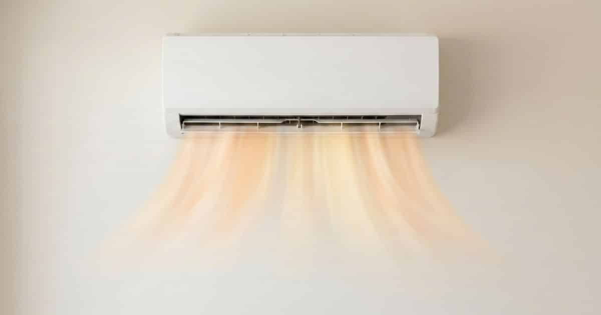 Witte split airco binnenunit hoog aan een lichte muur met zichtbare warme oranje luchtstroom die naar beneden stroomt uit de geopende lamellen