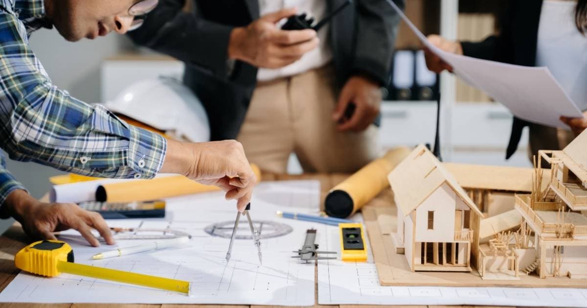 Een architect met een bril en een groen, wit en blauw geruit hemd gebruikt een passer om iets aan te duiden op een bouwtekening. Voor hem staat een houten maquette van een te renoveren gebouw. Op het plan liggen verschillende tekentools en een uitrolbaar meetlint.