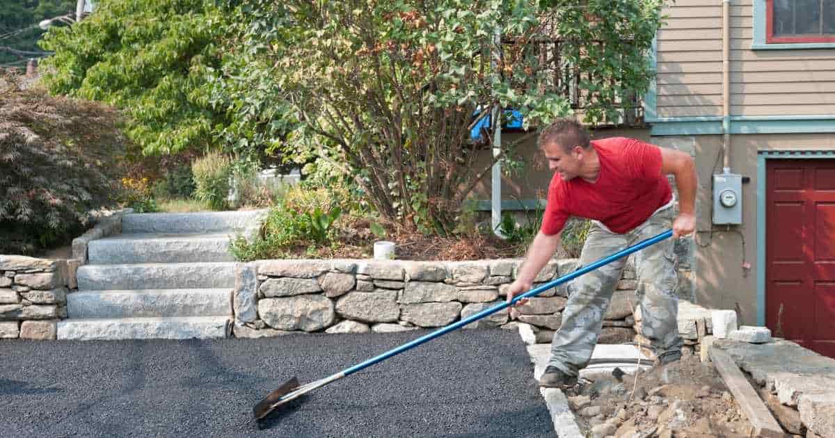 Een vakspecialist in rood t-shirt en grijze werkbroek legt een oprit met grind of split aan. De oprit geeft uit op een trapje dat naar een terras leidt.