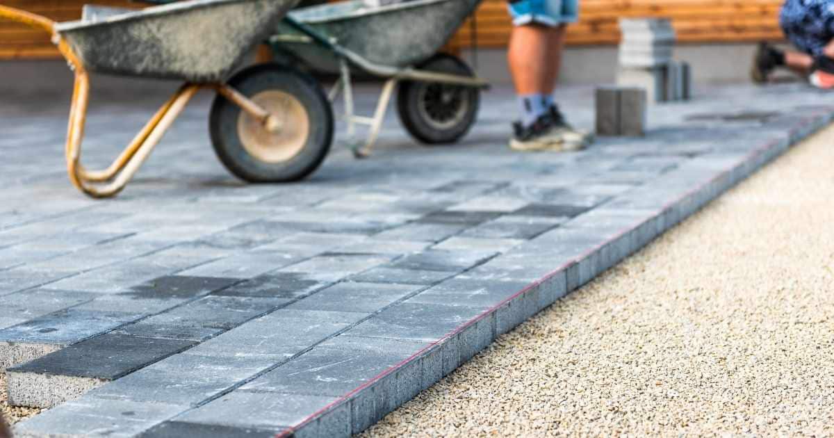 Een nieuwe oprit wordt aangelegd: op de laag stabilisé komt een laag grijze betonklinkers. In de verte twee vakmannen aan het werk, met elk een eigen kruiwagen met klinkers.
