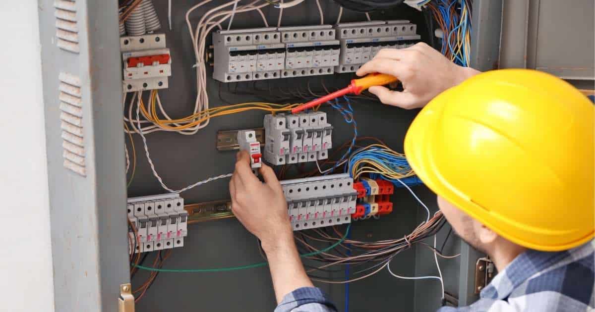 Een aannemer met gele helm werkt aan een werfkast. Hij gebruikt een schroevendraaier en draagt een wit met blauw geruit hemd.