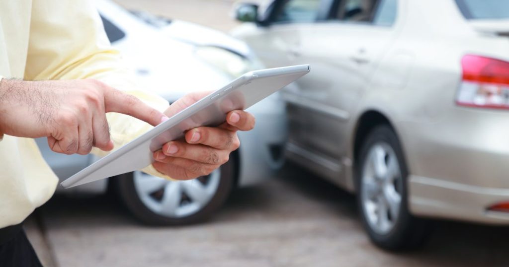 Een man voert de details van een kleine botsing in op een kleine tablet. Op de achtergrond twee auto's die zonet licht op elkaar ingereden zijn.