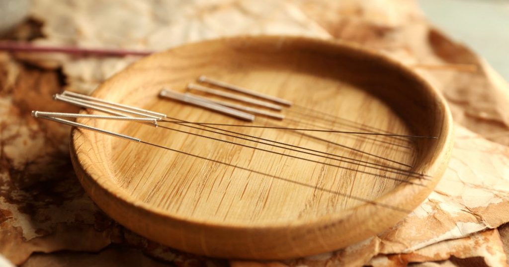 Verschillende naalden gebruikt voor acupunctuur liggen op de rand van een houten schaaltje. Het schaaltje staat op gekreukt papier in natuurlijke kleuren.