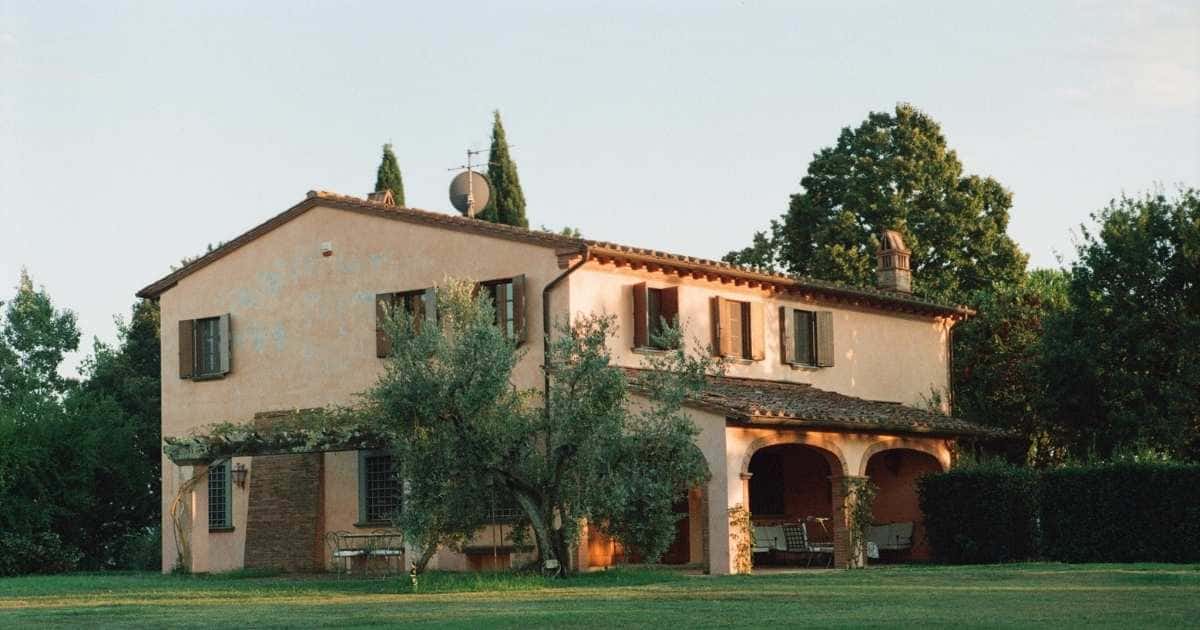 Een villa in zuiders design, met een oranjeroze gevel en keramische dakpannen. De villa staat op een grasveld omgeven met hoge bomen. Op het dak staat een satelliet.