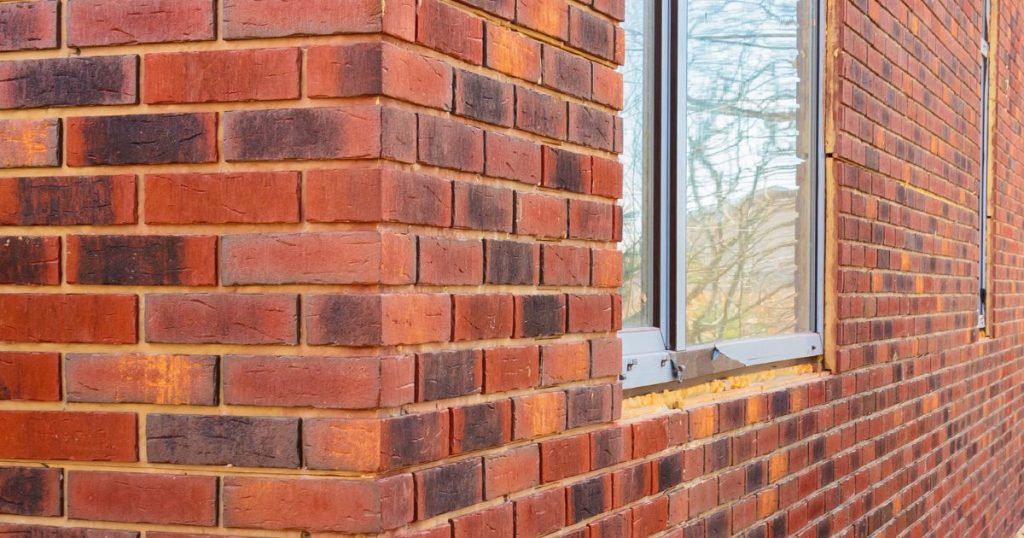 Close-up van een afgewerkte buitenmuur met rode baksteensteenstrips rond een raam, na plaatsing van de gevelafwerking.